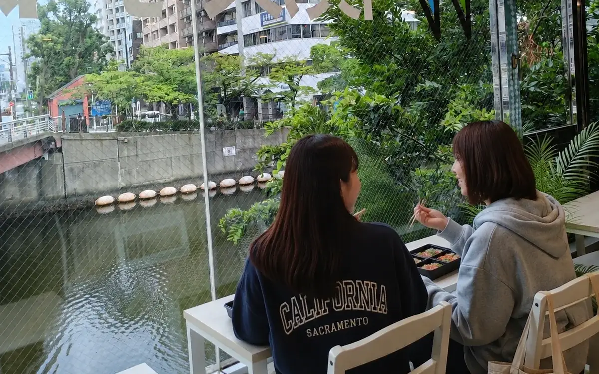 窓の外の川と緑を眺めながら、ラウンジでお弁当を食べる生徒たち